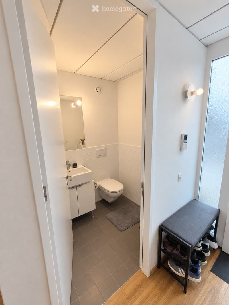 Modern bathroom with white tiles, a sink, toilet, mirror, and a small cabinet. A small bench with shoes is visible in the adjacent hallway.