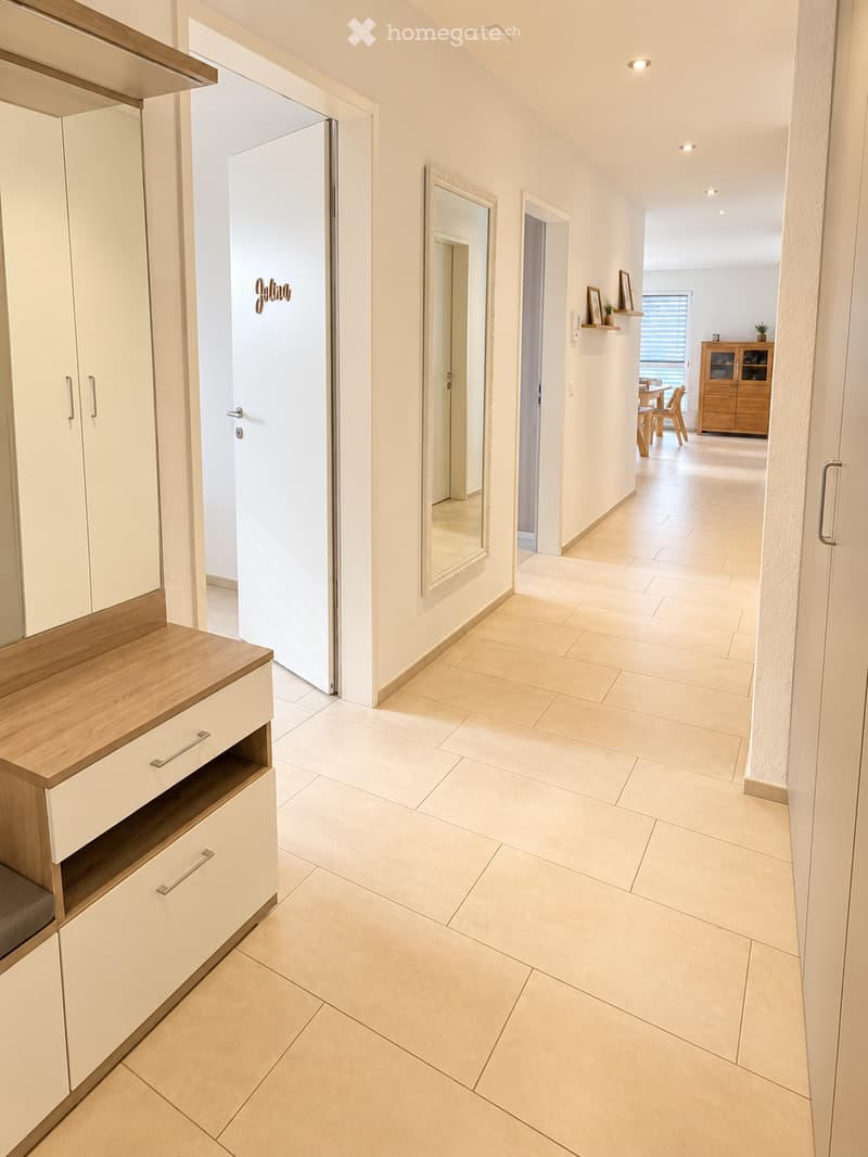 Modern hallway with beige tiled flooring, white walls, built-in storage unit with wooden countertop, and a mirror on the wall. The hallway leads to a dining area with a table and chairs visible in the background.