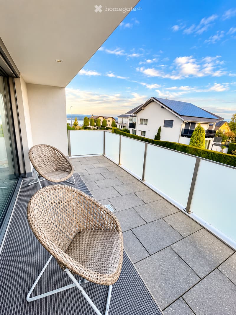 Balcony with two wicker chairs, modern glass railing, and a scenic view of the neighborhood and lake.