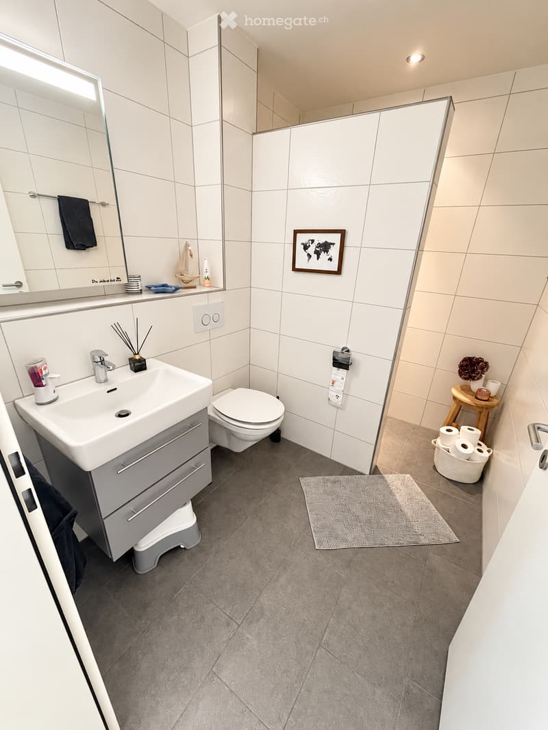Modern bathroom with white tiles, a sink with a mirror, a toilet, a small wooden stool, a picture frame, and a bath mat on the floor.