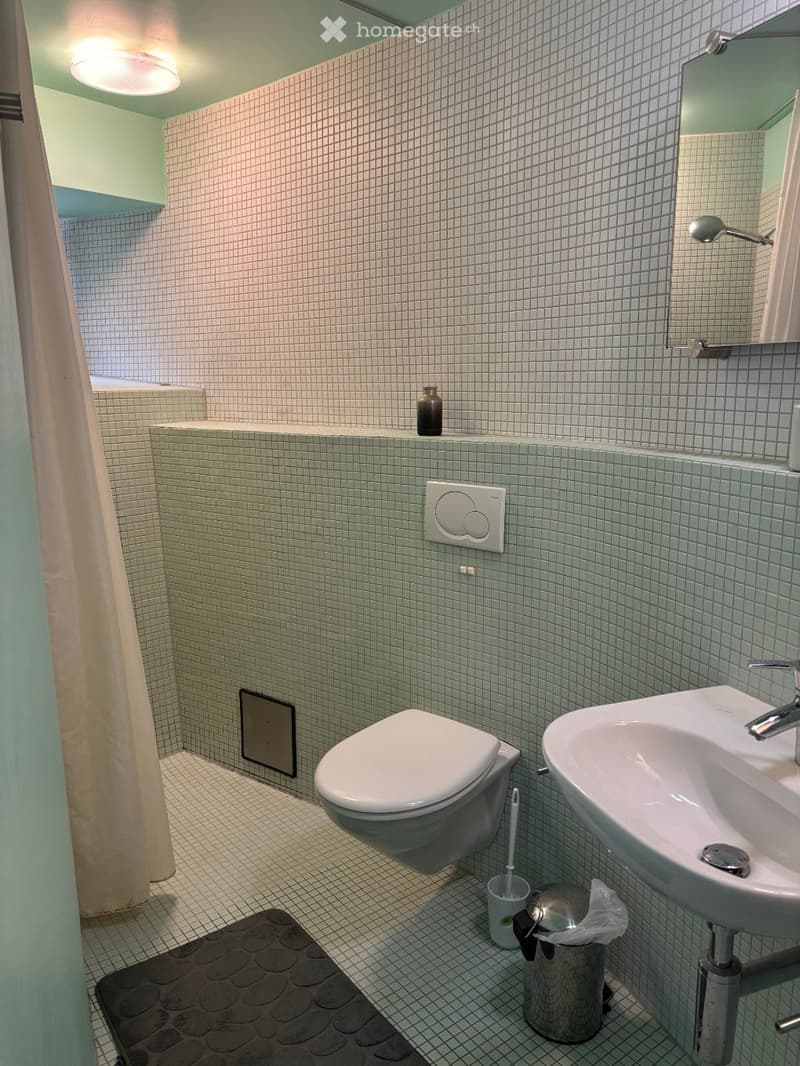 Modern bathroom with white tiled walls and floor, featuring a toilet, sink with chrome faucet, mirror, and a small decorative vase on the shelf.