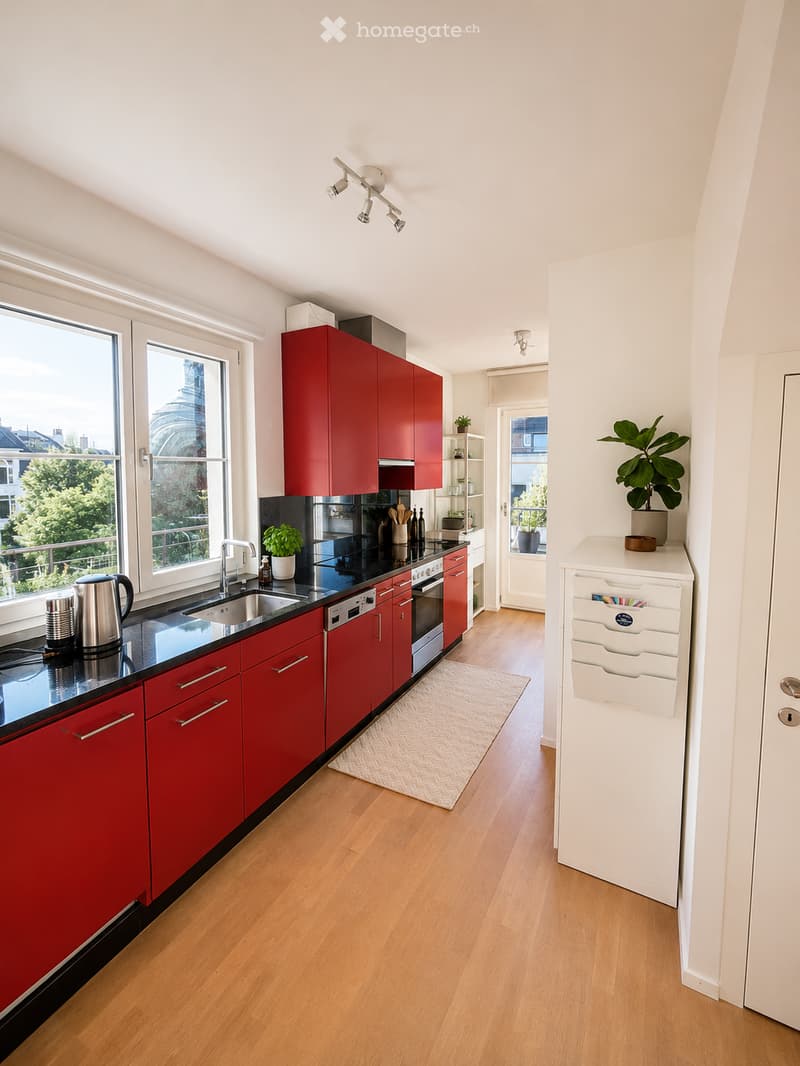 Modern kitchen with red cabinets, black countertops, stainless steel appliances, and wooden flooring. Features include a sink, oven, dishwasher, refrigerator, and various kitchen items on the counter.