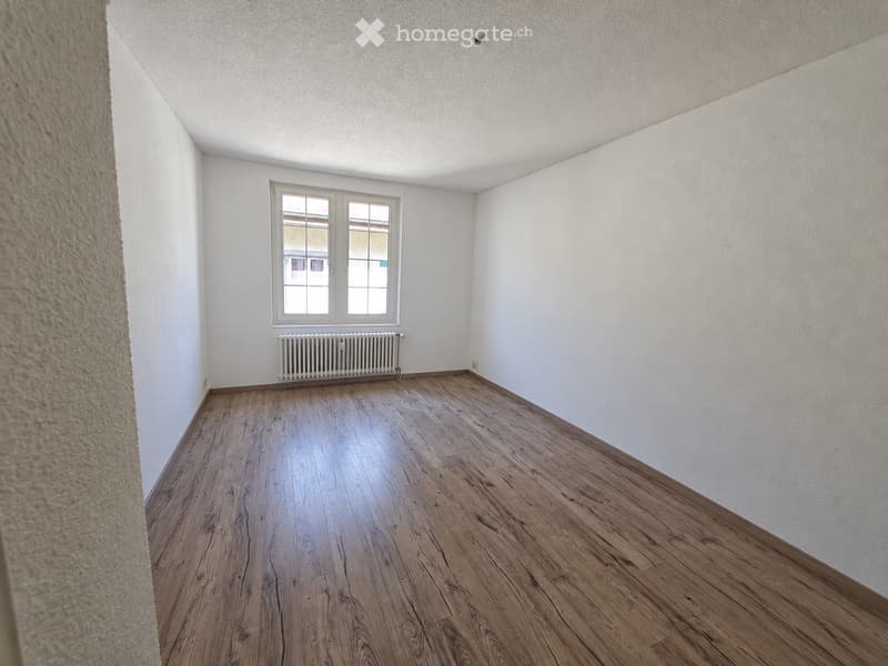Empty room with wooden flooring, white walls, and a window with a radiator below it.