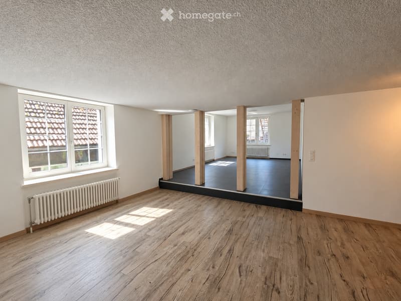 Empty room with wooden flooring, white walls, and large windows. The room features a radiator and a view of another room with dark flooring and wooden pillars.