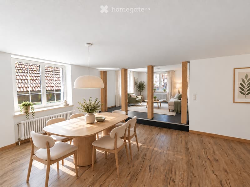 Bright dining area with a round wooden table, six chairs, a vase with flowers, and a bowl. The room features wooden flooring, large windows with a view of a tiled roof, and a pendant light. In the background, a living area with a sofa, coffee table, and a