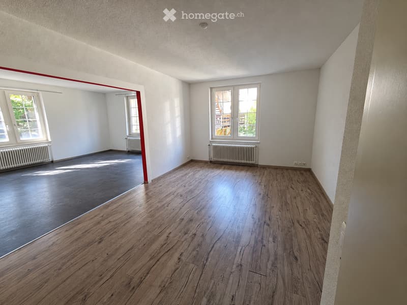 Empty room with wooden flooring, white walls, and large windows allowing natural light. Radiators are visible along the walls.