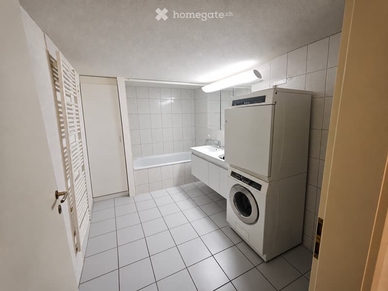Modern bathroom with white tiled walls and floor, featuring a bathtub, sink, and a stacked washing machine/dryer unit.