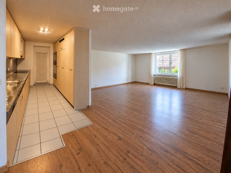 Empty room with wooden flooring, white walls, and a large window with curtains. The room is connected to a kitchen area with wooden cabinets, a sink, and a microwave.
