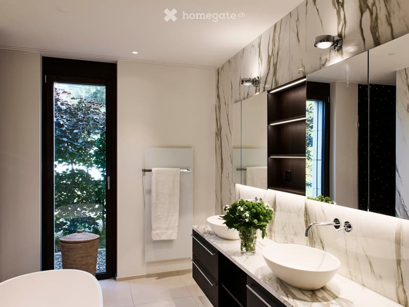 Modern bathroom with marble walls, double sink vanity, large mirror, bathtub, and a view of greenery through a large window.