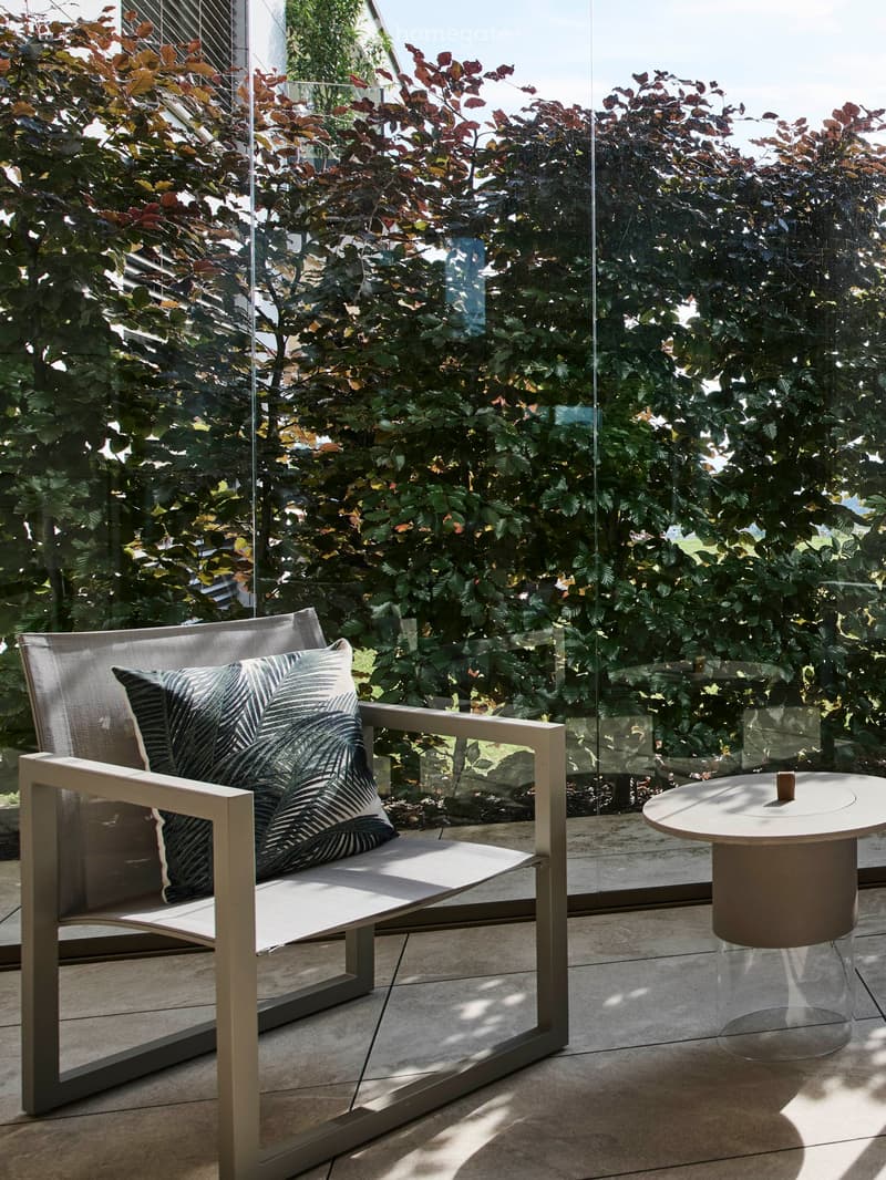 Modern outdoor seating area with a single chair, cushion, and a round table, enclosed by a glass railing with lush greenery visible beyond.