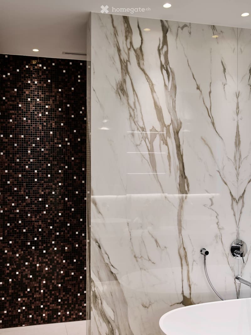 Modern bathroom with marble wall tiles, dark mosaic tiles on one wall, and a bathtub with a chrome faucet.
