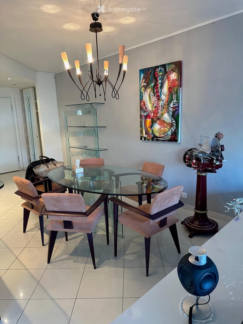 Modern dining area with a glass table, six upholstered chairs, a chandelier, a colorful painting, and a speaker on a side table.
