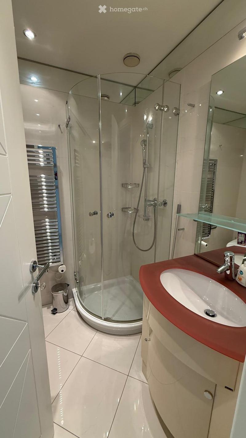 Modern bathroom with a glass-enclosed shower, a sink with a red countertop, a mirror, and a toilet paper holder. The floor is tiled in white, and the walls are light-colored.