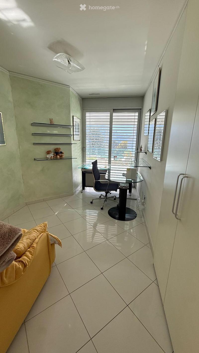 Bright, modern room with tiled flooring, a comfortable sofa, a desk with a chair, shelves with decorative items, and large windows offering a view.