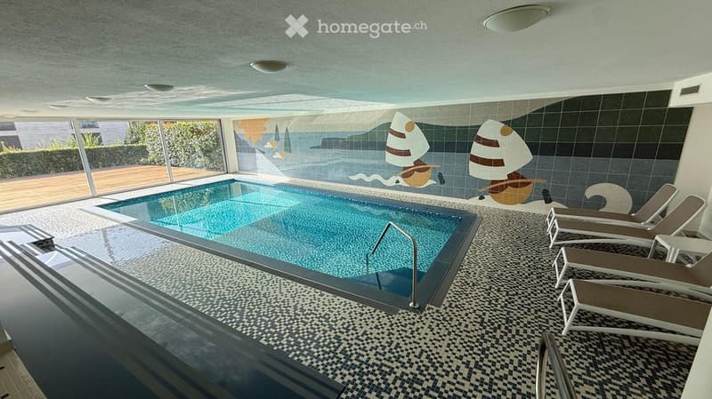 Indoor swimming pool area with lounge chairs, decorative wall art, and large windows overlooking an outdoor patio.