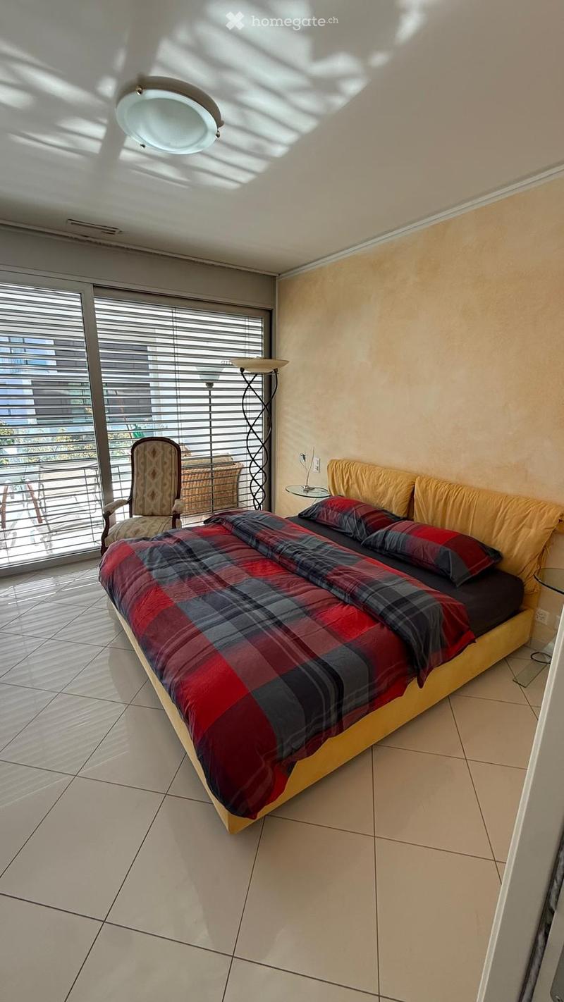 Bright bedroom with a large bed, plaid duvet, and matching pillows. A wicker chair and small table are near the window, which has blinds and offers a view of the balcony.