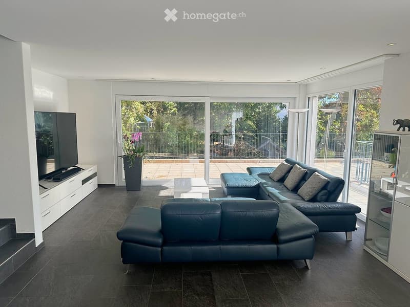 Modern living room with dark grey leather sofa, coffee table, flat-screen TV on white cabinet, and large glass doors leading to a balcony.