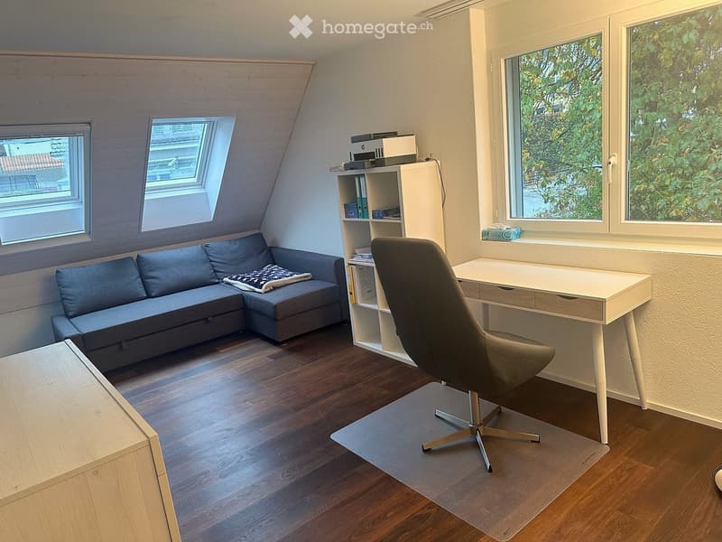 Cozy attic room with wooden flooring, large windows, a grey sectional sofa, a desk with a chair, and a shelf with books and a printer.
