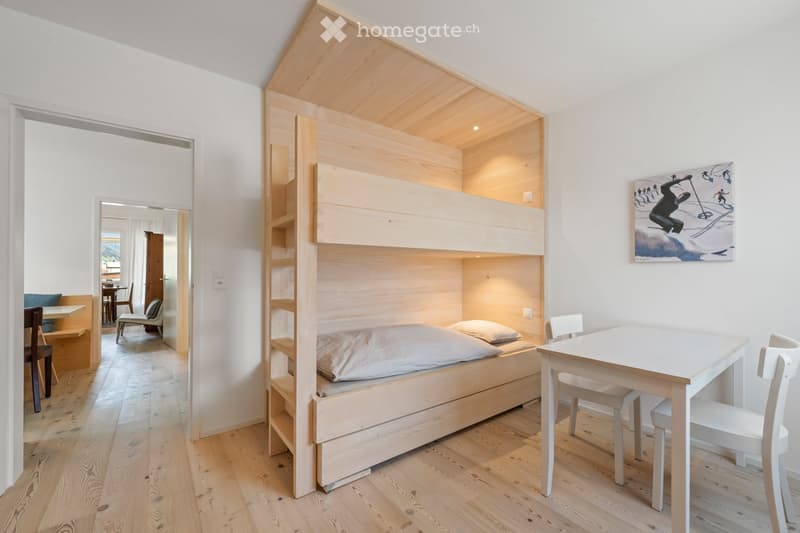 Cozy room with a bunk bed, small table, chairs, and a picture on the wall. The room has wooden flooring and white walls.