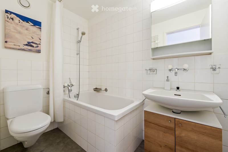 Modern bathroom with white tiles, a toilet, a bathtub, a sink with a wooden cabinet, a mirror, and a showerhead.