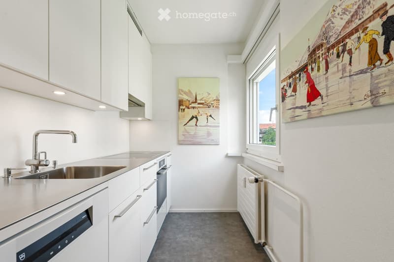 Modern kitchen with white cabinets, stainless steel sink, oven, and two large windows. Two paintings adorn the walls.