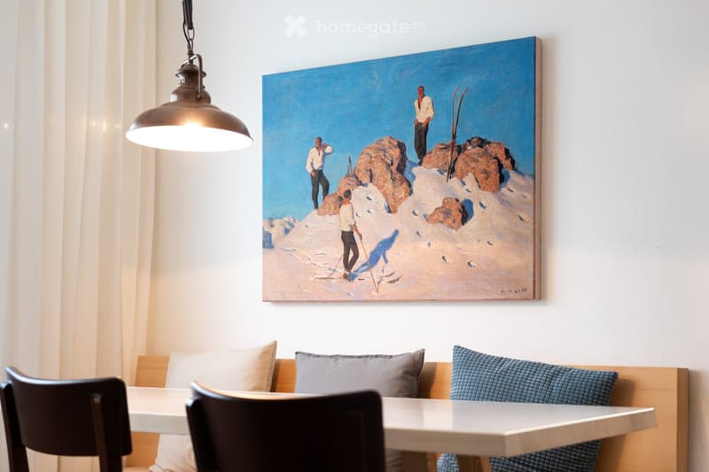 A modern dining area with a white table, black chairs, a wooden bench with cushions, and a large painting of skiers on a snowy mountain above the seating area.