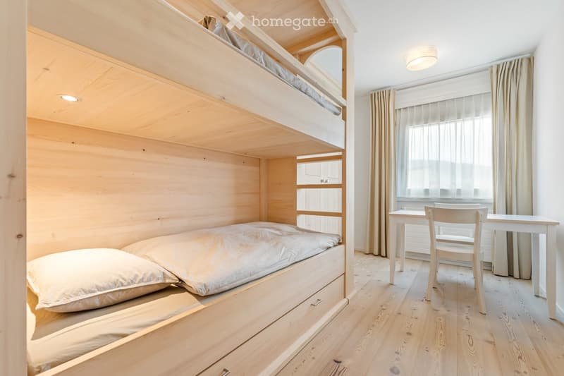 Cozy bedroom with a bunk bed, a small table, and a chair, featuring wooden walls and flooring.