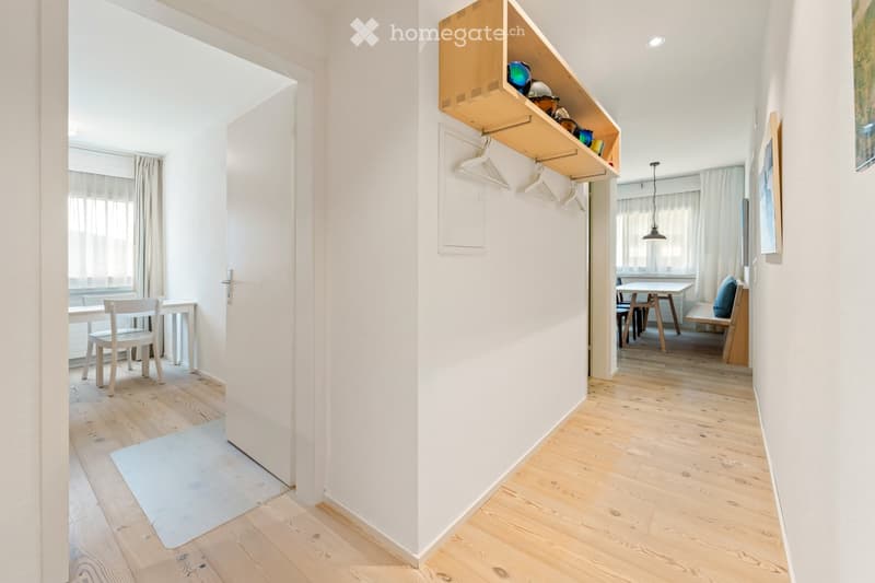 A bright, modern hallway with wooden flooring, white walls, and built-in shelves holding decorative items. The hallway leads to a dining area with a table and chairs, and a bench with a cushioned seat is visible in the background.