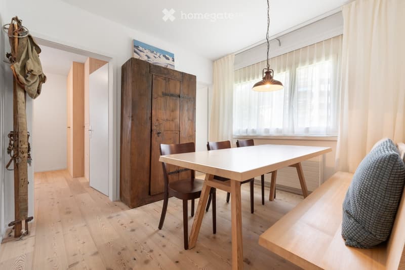 Bright dining area with a modern white dining table, wooden chairs, a wooden bench with a cushion, and a large wooden cabinet. Natural light streams in from large windows with white curtains.
