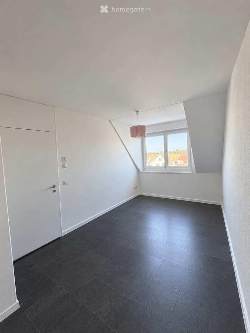 Empty room with dark grey tiled floor, white walls, and a large window offering a view of houses outside. A single hanging light fixture is visible.