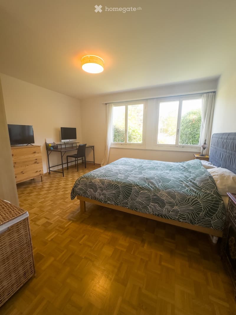 Well-lit bedroom with wooden flooring, large window with white curtains, bed with patterned duvet, desk with chair and computer monitor, nightstand with lamp, and a wicker basket on the floor.