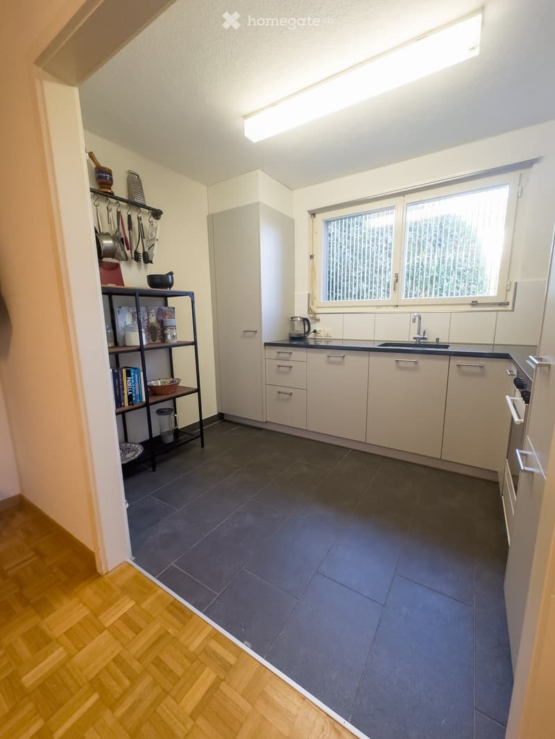 Modern kitchen with white cabinets, dark grey tiles, and a window above the sink. A shelf holds various kitchen items and books.