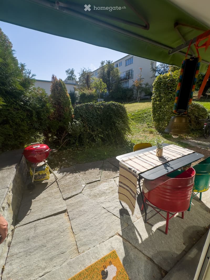 Outdoor patio with stone flooring, a table with chairs, a red barbecue, and a small potted plant. Greenery and a building are visible in the background.