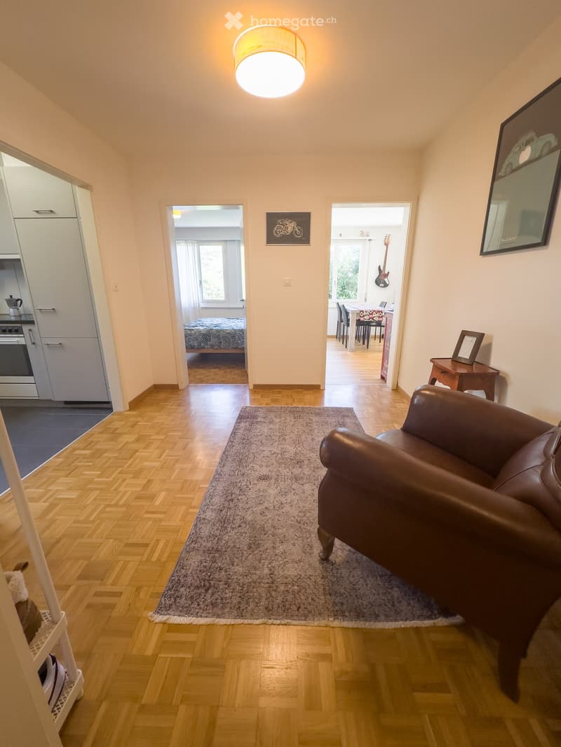 A modern living room with a brown leather sofa, a grey rug, a small wooden table, and a guitar in the adjacent room. The space features wooden flooring and white walls.