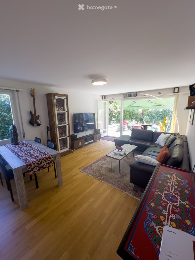 Modern living room with wooden flooring, large windows, a dark grey sectional sofa, coffee table, dining table, and a TV stand. Features a guitar on the wall and a colorful rug.