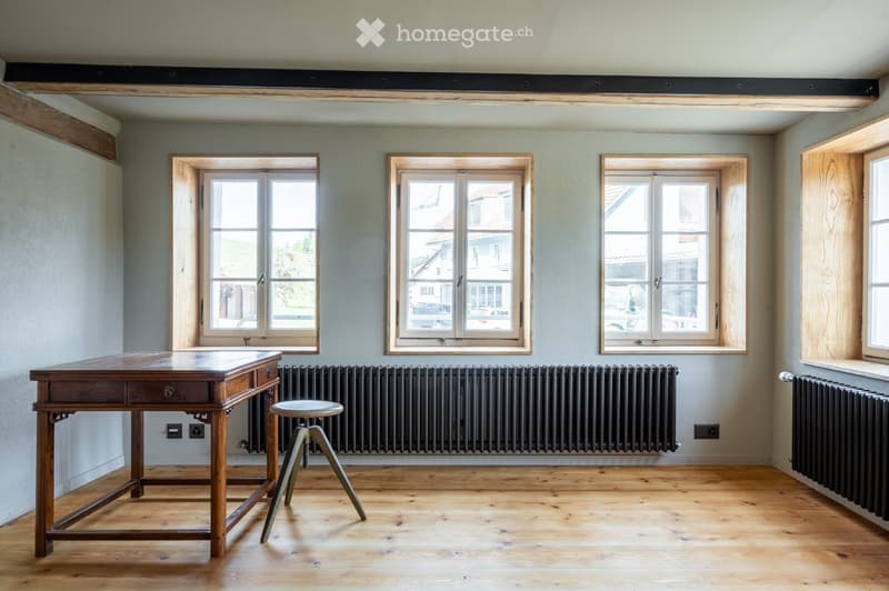 Bright, unfurnished room with wooden flooring, large windows, radiators, and a wooden table with a stool.
