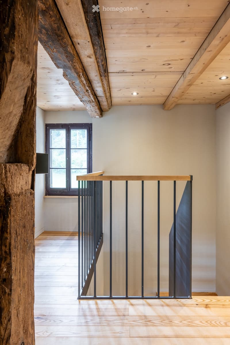 Spacious hallway with wooden floors, exposed wooden beams, and a modern metal railing leading to a window with a view of greenery.
