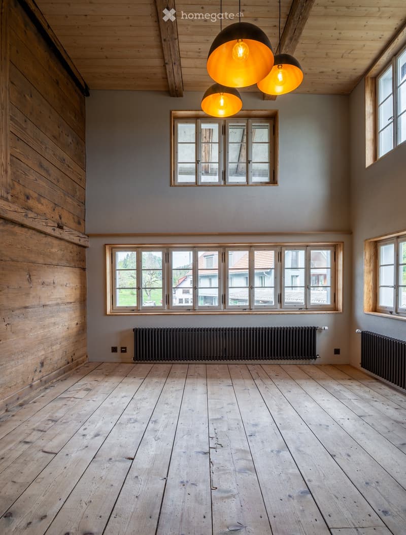 Empty room with wooden floors, exposed wooden beams on the ceiling, large windows, and modern pendant lights.