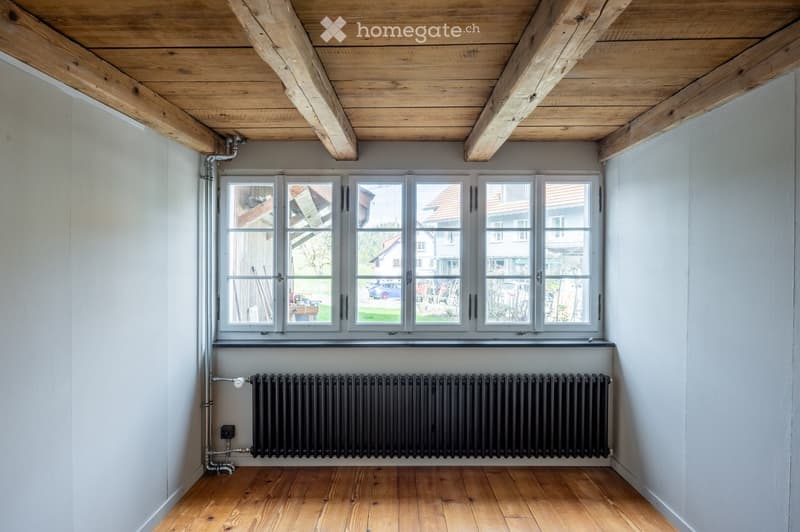 Empty room with wooden floor, white walls, large windows, radiator, and exposed wooden ceiling beams.