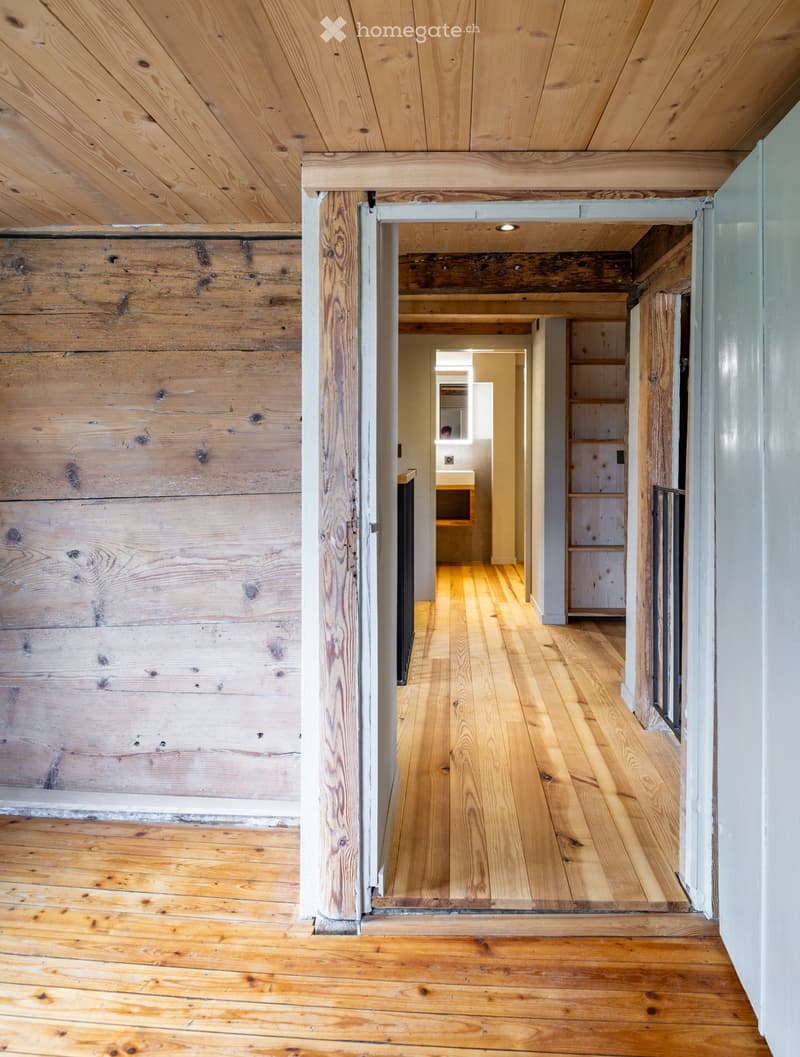 A long, narrow hallway with wooden floors and exposed wooden beams on the ceiling. The walls are made of reclaimed wood planks, and the hallway leads to a small bathroom with a sink and mirror visible at the end.