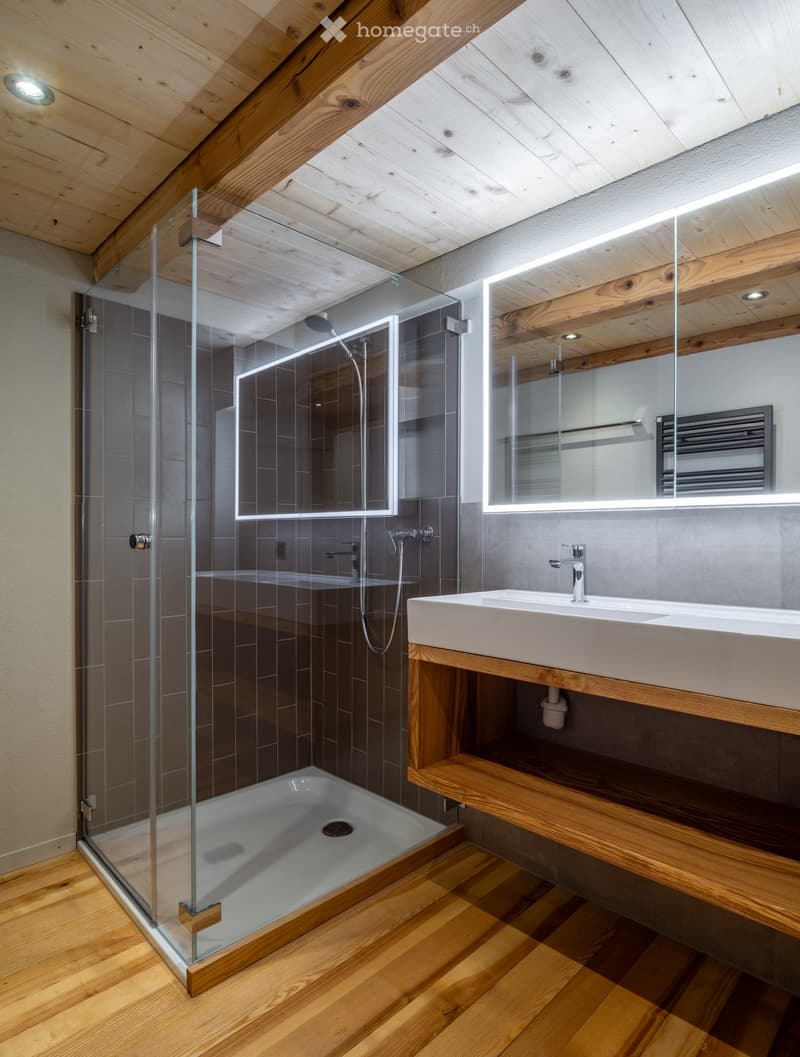 Modern bathroom with wooden ceiling beams, glass shower enclosure, white sink, and built-in mirror with LED lighting.