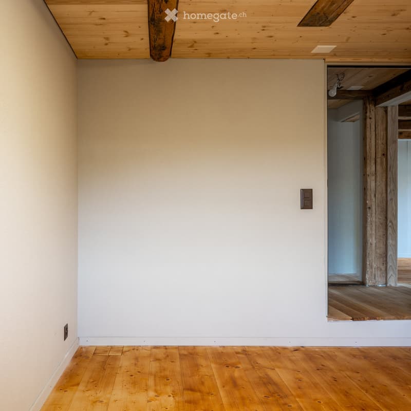 Empty room with light-colored walls and wooden flooring, featuring a simple wooden ceiling and visible electrical outlets.