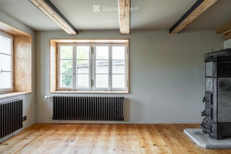 Empty room with wooden flooring, exposed wooden beams on the ceiling, large windows, radiators, and a stone fireplace base.