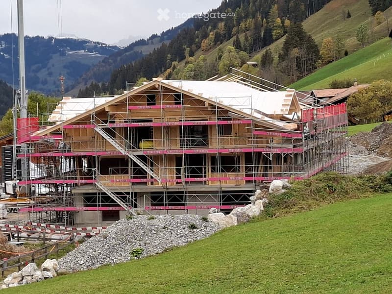 A large wooden building under construction, surrounded by scaffolding and safety barriers, set in a green, mountainous landscape.
