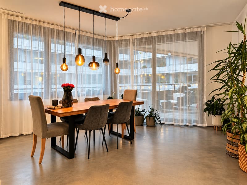 Modern dining area with a wooden table, grey upholstered chairs, a vase with red flowers, and several potted plants near large windows with sheer curtains.