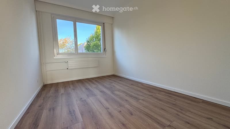 Empty room with wooden flooring, white walls, and a large window with a view of trees and a building outside.