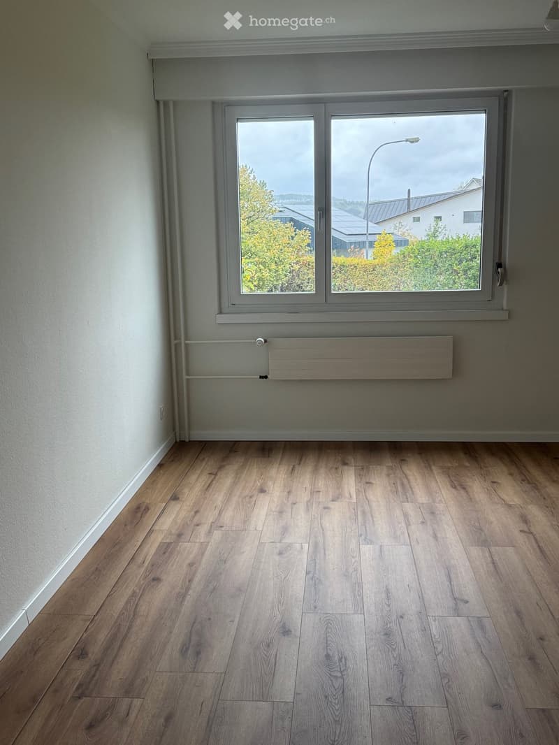 Empty room with light wooden flooring, white walls, and a large window offering a view of greenery and buildings outside.