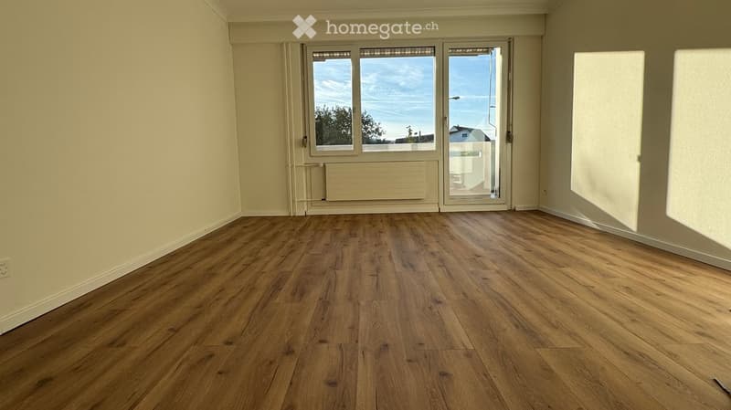 Empty room with wooden flooring, large windows, and a sliding glass door leading outside.