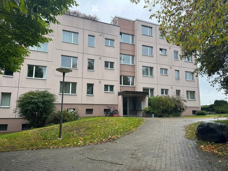 A multi-story residential building with light pink walls and numerous windows, surrounded by greenery and a paved pathway.