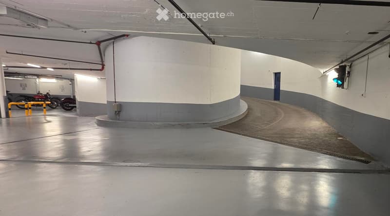 Underground parking area with concrete floors and walls, featuring a curved central pillar, a few parked motorcycles, and yellow safety barriers.